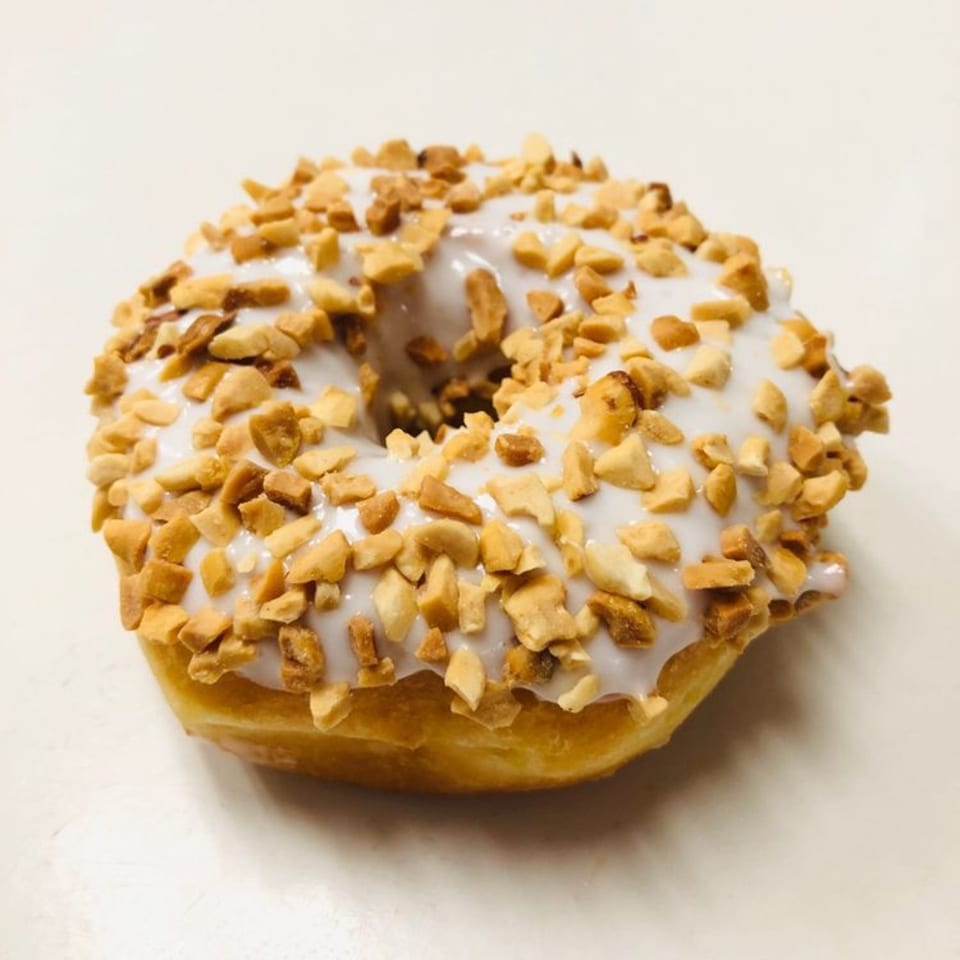 A Donut Taco Palace donut with white icing and chopped peanuts on a white surface.