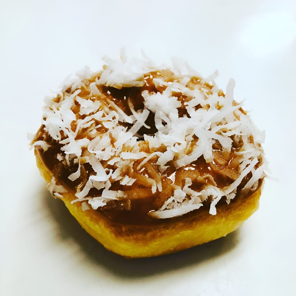 A Donut Taco Palace donut with caramel frosting and shredded coconut on a white background.