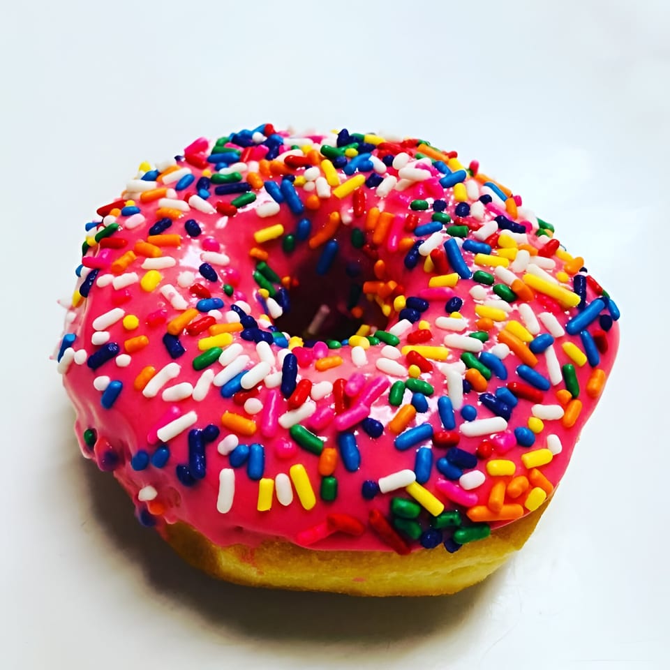 A Donut Taco Palace donut with bright pink icing and colorful sprinkles on a white background.